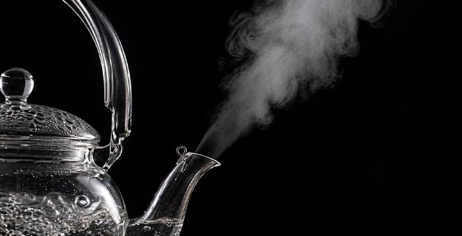 Glass tea kettle releasing visible vapor against a dark background